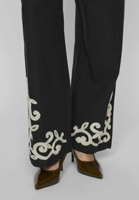Pantalones negros con patrones de remolinos bordados en crema en el dobladillo, emparejados con zapatos de tacón marrón y puntera fina. La tela parece suave y tejida.