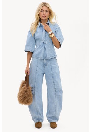 Femme blonde portant une chemise en jean bleu clair et un jean à jambes larges, tenant un sac à main marron duveteux et portant des chaussures marron sans lacets.