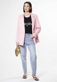 Blazer rosa de doble botonadura sobre una camiseta negra con letras metálicas, combinado con jeans azul claro y un clutch dorado. Sandalias negras de tiras.