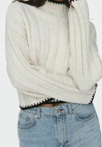 Personne portant un pull crème côtelé avec des bordures noires et un jean taille haute bleu clair, les bras croisés sur la poitrine.