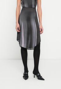 Asymetrická čierna saténová sukňa s hladkou textúrou, s vysokým a nízkym lemom, v kombinácii s čiernymi nepriehľadnými pančuchami a lodičkami so špicatou špičkou.