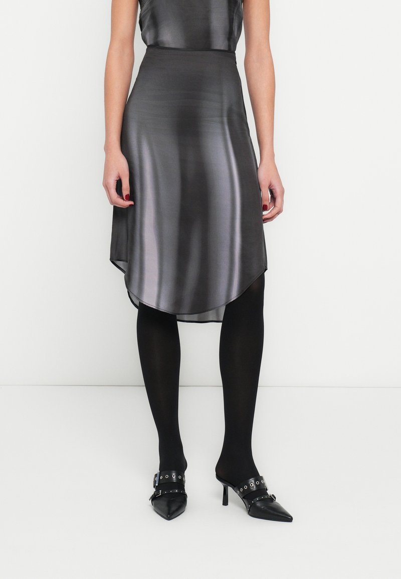 Asymetrická čierna saténová sukňa s hladkou textúrou, s vysokým a nízkym lemom, v kombinácii s čiernymi nepriehľadnými pančuchami a lodičkami so špicatou špičkou.