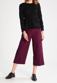 Zwarte, gegoteerde langemouwen top met bloemenontwerp, gecombineerd met brede, cropped bordeauxrode broek en zwarte platte schoenen.