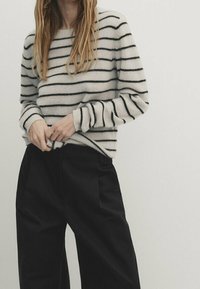 Frau mit beigem Pullover mit schwarzen horizontalen Streifen und schwarzen weit geschnittenen Hosen, die den unteren Teil des Pullovers anpasst.