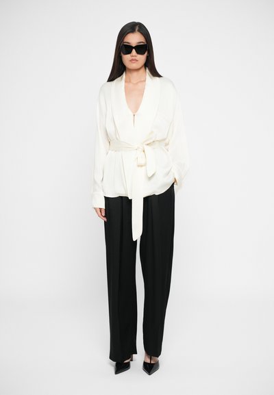 Blusa avvolgente color crema con scollatura a V profonda, vita legata e maniche fluttuanti, abbinata a pantaloni neri a gamba larga e tacchi neri appuntiti.