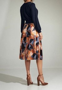 Femme portant un blouse noire à manches longues, une jupe midi à motif géométrique et des chaussures à talons hauts brun avec brides à la cheville, se tenant sur le côté.