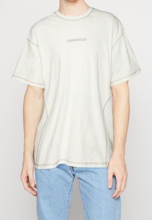 Homme portant un t-shirt blanc ample avec le petit texte "LETS FRANS" sur la poitrine et un jean bleu clair, debout devant un fond uni.