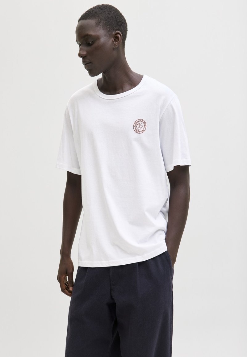 Ung mand iført en hvid T-shirt med et lille rundt emblem på brystet og mørke bukser, kigger ned med den ene hånd i lommen.