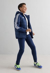 Granatowa kurtka ocieplana w białe paski, noszona na białej koszuli i granatowych legginsach, zestawiona z niebieskimi i zielonymi butami sportowymi.