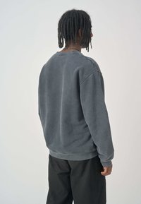 Grauer Rundhalsausschnitt-Pullover aus weichem Material, mit lockerer Passform, gerippten Bündchen und Saum sowie einem schlichten Rückendesign. Kombiniert mit schwarzen Hosen.