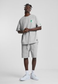 Grå bomulls t-skjorte med grønn logo, kombinert med grå shorts og hvite joggesko. Har en avslappet passform og myk tekstur.