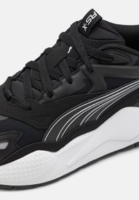 Chaussure de sport noire avec tige en tissu texturé, lacets ton sur ton et semelle en caoutchouc blanche ornée d'un motif à points et d'accents argentés.