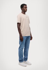 T-shirt beige in cotone con maniche corte e un logo sul petto, abbinata a jeans dritti azzurri e sneakers bianche.