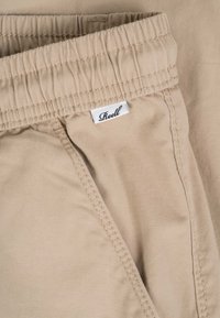 Pantalones con cinturilla elástica beige con etiqueta visible de la marca Reell y costuras detalladas a lo largo del borde del bolsillo.