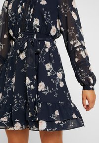 Mörkblommig klänning med långa genomskinliga ärmar och knytning i midjan. Tyget har rosa och vita blommor mot en marinblå bakgrund.