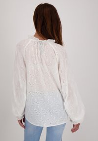 Witte blouse met een zigzag uitsnijdpatroon, gerimpelde halslijn en zwierige mouwen, gecombineerd met lichtblauwe jeans.