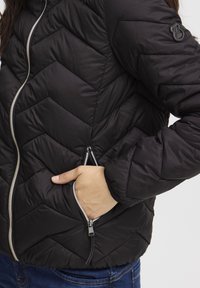 Črna puhovka z vzorcem cik-cak, srebrnim zadrgo in stranskimi žepi. Mehka, sijoča tkanina z rebrastimi manšetami.