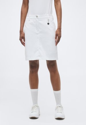 Persoană purtând o fustă albă până la genunchi cu bucle pentru curea, nasture frontal, buzunare laterale, șosete albe și adidași albi pe un fundal simplu.