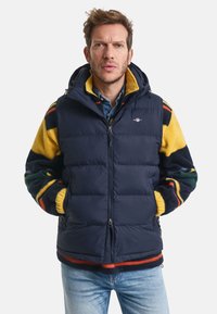 Navy Puffer-Weste mit Ärmeln aus gelbem, grünem und rotem gemustertem Fleece, Reißverschluss, verstellbarer Kapuze und zwei Vorderaschen.