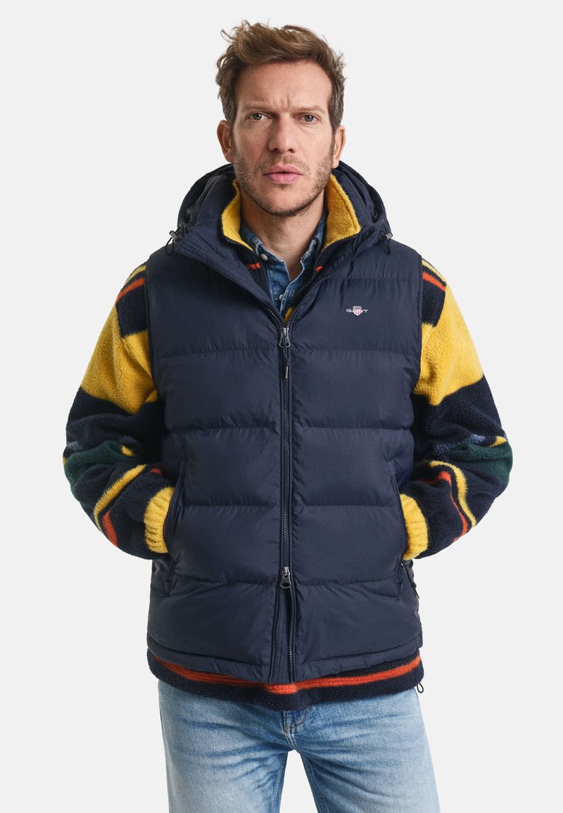 Navy Puffer-Weste mit Ärmeln aus gelbem, grünem und rotem gemustertem Fleece, Reißverschluss, verstellbarer Kapuze und zwei Vorderaschen.