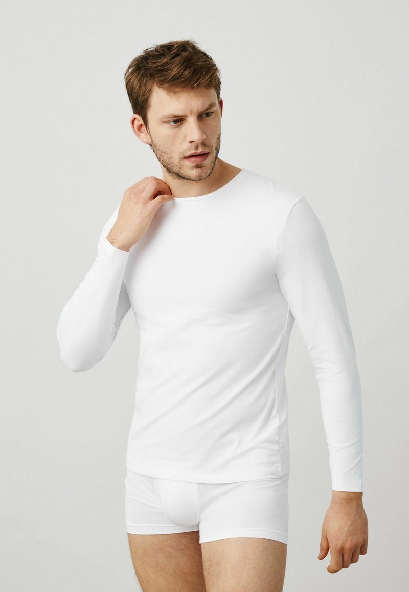 Camisa blanca de manga larga hecha de tela suave, diseño ajustado, escote redondo. Combinada con pantalones cortos blancos, construcción sin costuras, líneas limpias.