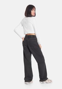 Kobieta o prostych ciemnych włosach, ubrana w biały, krótki top z długim rękawem, luźne czarne dżinsy i białe trampki, stojąca na białym tle.