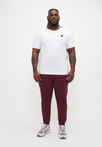 Hvit t-skjorte med korte ermer og en liten svart logo, sammen med burgunder tettsittende bukser og grå joggesko med en kraftig design.