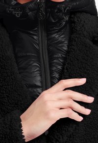 Zwarte fleece buitenjas met een ritssluiting en een glanzende zwarte puffer binnenvoering. Heeft een hoge kraag en gegolfde manchetten, en toont een handpositie.