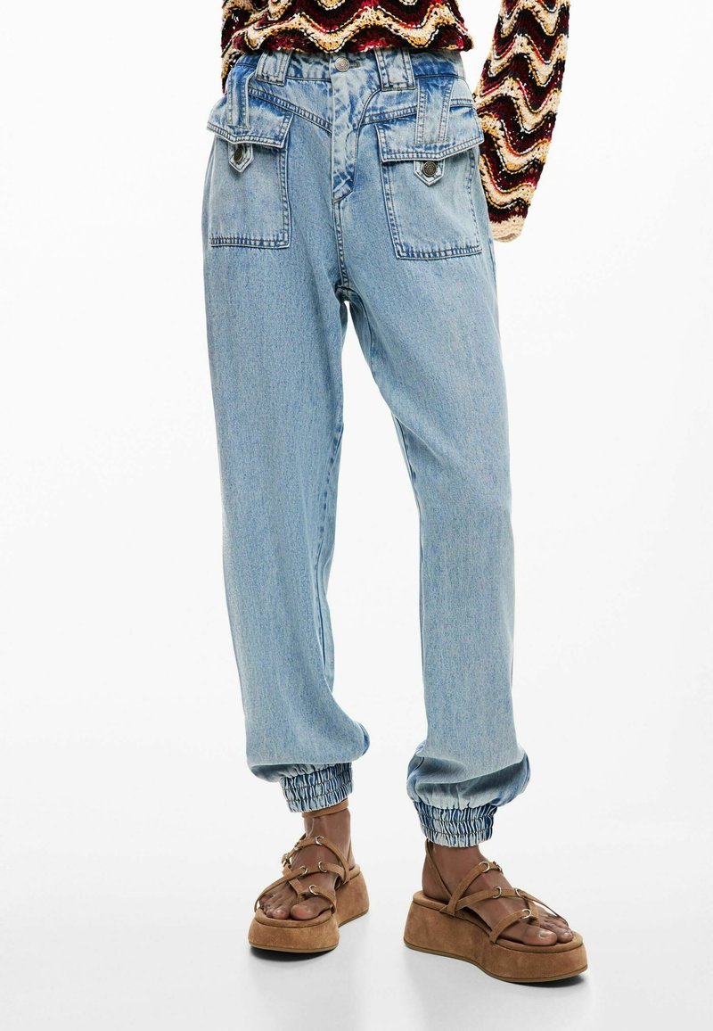 Hellblaue Jeans-Jogginghosen mit elastischen Bündchen und großen vorderen Klappentaschen, kombiniert mit braunen Plateausandalen und einem mehrfarbigen Stricktop.