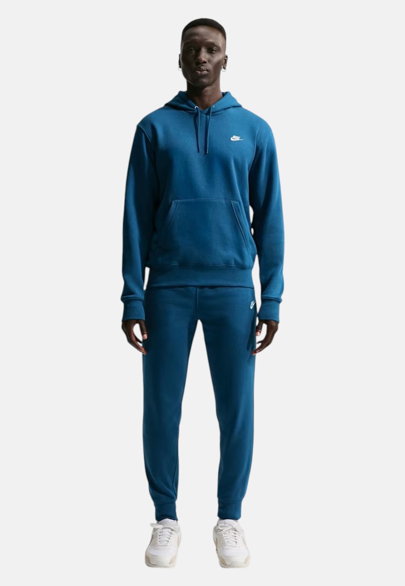 Homme portant un sweat à capuche Nike bleu et un pantalon de jogging assorti avec des baskets blanches, debout devant un fond blanc uni.