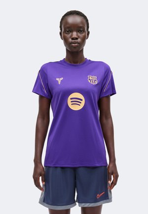 Femme portant un maillot de football violet du FC Barcelone avec des logos dorés et un short Nike bleu foncé, debout devant un fond uni.