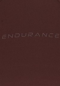 Mörkröd idrottstopp med ribbad textur. Har upphöjd text "ENDURANCE" över bröstet i en matchande färg. Enkel, figurnära design.