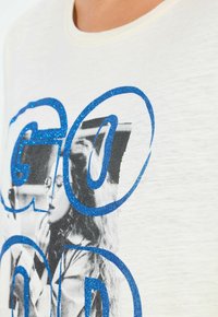 Primer plano de una camiseta blanca con texto azul brillante superpuesto a una foto en blanco y negro de dos mujeres cepillándose los dientes.