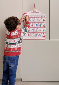 Calendar de Advent fabricat din țesătură, cu buzunare numerotate, având designuri colorate de sărbătoare. Un copil îmbrăcat într-un pulover cu model adaugă o decorațiune aproape de vârf.