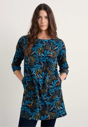 Tunique imprimée florale en bleu foncé, avec des motifs de feuilles en teal, or et crème. Les manches longues et les poches latérales renforcent la fonctionnalité.