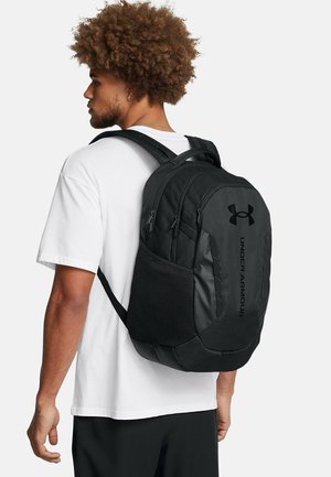 Juoda kuprinė iš tvirtos medžiagos su paminkšintomis petnešomis, keliais užsegamais skyriais, akių šonine kišene ir „Under Armour“ logotipo detalėmis.