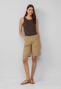 Frau mit braunem, ärmellosem Top, beigen, knielangen Shorts mit umgeschlagenen Säumen und Leopardenmuster-Ballettschuhen, die vor einer schlichten Wand steht.