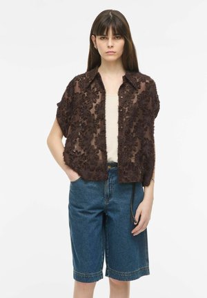 Femeie purtând o bluză texturată maro închis, transparentă, cu mâneci scurte peste un top alb, asortată cu pantaloni scurți din denim albastru până la genunchi.