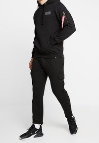 Felpa nera con tasca frontale, cappuccio regolabile e tasca laterale con zip. Abbinata a pantaloni cargo neri con tasca laterale. Sneakers nere.