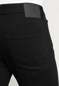 Black denim jeans feature a smooth texture, standard back pockets, and a leather patch with branding on the waistband.