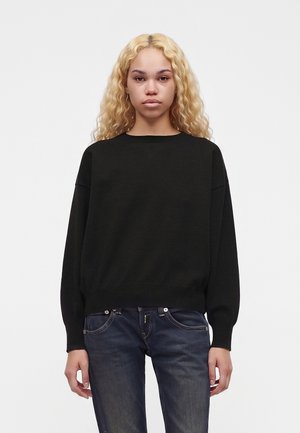 Ung person med langt krøllet blondt hår iført en sort sweater og mørkeblå jeans, stående mod en ensfarvet hvid baggrund.