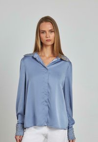 Blusa de seda azul con mangas largas, cierre frontal con botones y hombros y puños adornados con remaches metálicos para un detalle adicional.