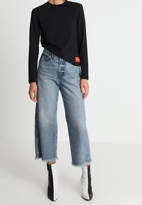 Chemise noire à manches longues, jeans bleu taille haute à jambes larges avec ourlet effiloché, associés à des bottines noires et blanches.