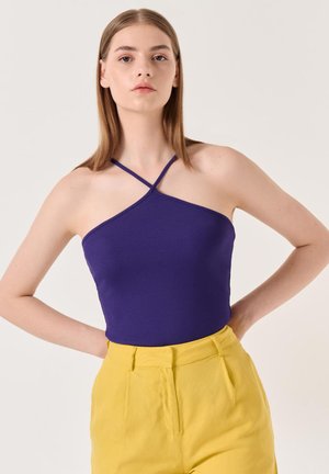 Top a halter blu porpora con design a fascia attorcigliata, abbinato a pantaloni gialli a vita alta, che mettono in risalto una silhouette aderente e colori vivaci.