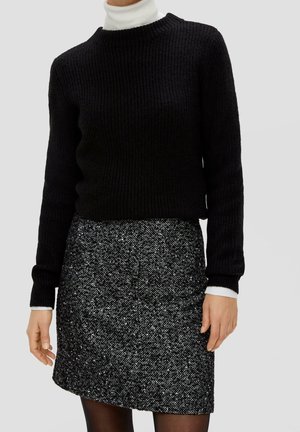 Femme portant un pull en maille noir par-dessus un col roulé blanc, accompagné d'une jupe droite texturée noire et blanche et de collants noirs.