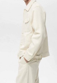 Giacca in denim color crema con colletto, tasche frontali e vestibilità rilassata. Presenta chiusure con bottoni e una texture morbida.