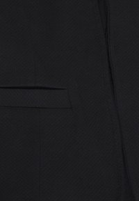 Svart tyg närbild med en strukturerad väv. Synlig horisontell söm med en liten, platt ficka ovanför. Dragkedjedetalj på sidan.