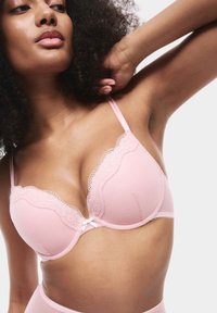 Žena s kudrnatými vlasy, oblečená v lehkém růžovém podprsence s krajkovým lemem, zvedá jednu ruku za hlavu na jednobarevném pozadí.