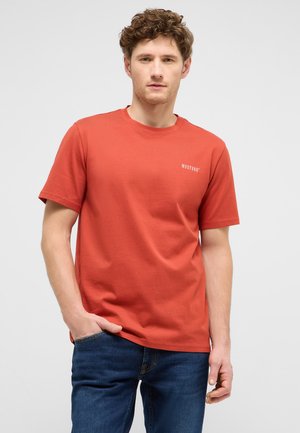 Young man with curly hair wearing a red Mustang T-shirt and blue jeans, standing with one hand in his pocket against a plain background.