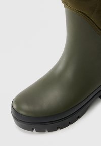 Groene rubberen regenlaarzen met een zwarte getailleerde zool en gequilt stofdetail rond de enkel.